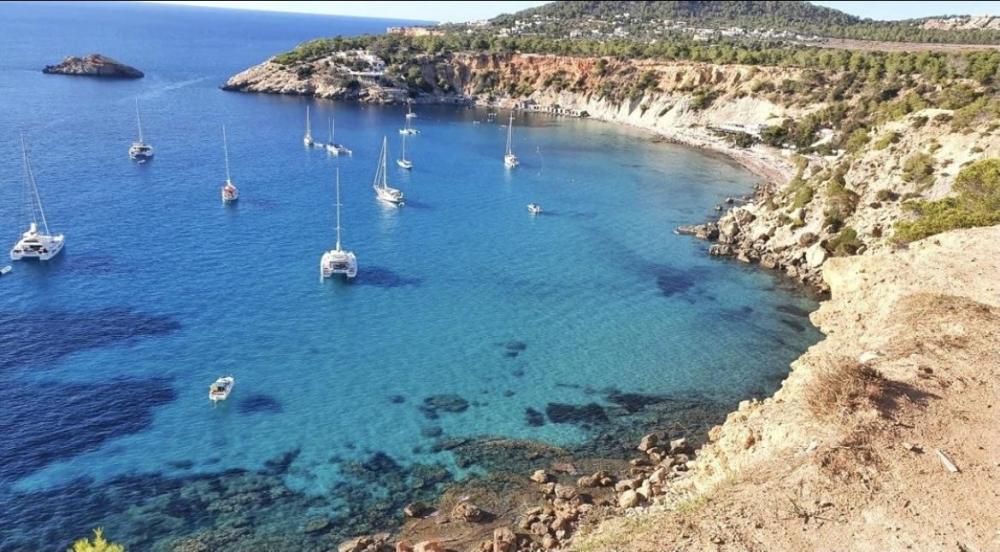 A rocky coastal cove with clear blue water and several anchored sailboats.