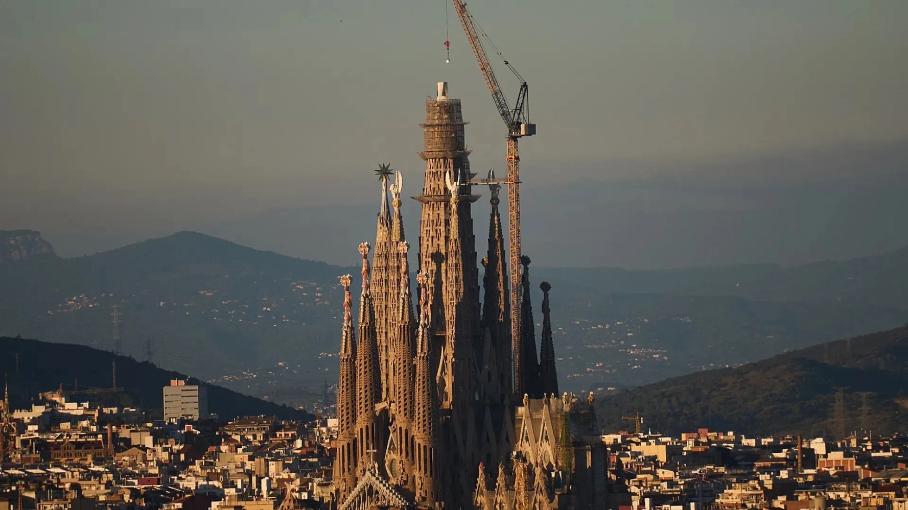 Aerial view of a large Gothic cathedral with cranes, surrounded by urban landscape and mountains.
